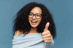 mi piace ritratto di donna felice con acconciatura afro, mostra il pollice in su, esprime un'opinione positiva, guarda da parte, indossa occhiali da vista e maglione, isolato su sfondo blu. persone, accordo, approvazione foto