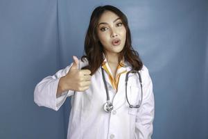 ritratto di una giovane donna asiatica medico, un medico sorride e mostra i pollici in su o il segno ok isolato su sfondo blu foto