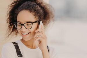 ritratto ravvicinato di donna allegra con capelli crespi, soddisfatta delle tariffe per le chiamate telefoniche, focalizzata verso il basso, tiene uno smartphone moderno, telefoni al migliore amico, discute le ultime notizie. concetto di tecnologia foto