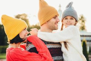 persone e concetto di relazione. la famiglia trascorre momenti indimenticabili insieme, si abbraccia, indossa cappelli lavorati a maglia alla moda. adorabile bambina con le trecce abbraccia il padre, guarda felicemente nella fotocamera foto