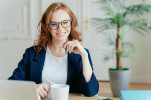 giovane imprenditrice di successo prepara il rapporto per il progetto di avvio, cerca informazioni sul computer portatile, si concentra sul suo compito, tiene una tazza di caffè, la penna tiene la mano sotto il mento pone al chiuso foto