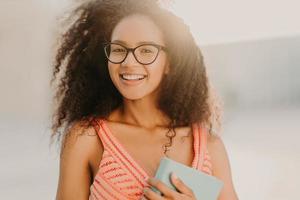 foto di uno studente afroamericano spensierato in abito estivo, tiene un libro, si prepara per l'esame, passeggia per le strade della città, si gode una calda giornata di sole, ha un sorriso affascinante sul viso. persone, concetto di etnia