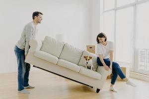 una coppia felice porta insieme un moderno divano bianco con il cane, posiziona i mobili in soggiorno, si preoccupa del miglioramento del design degli interni, inizia a vivere in una nuova casa, posa in un appartamento moderno, divertiti foto