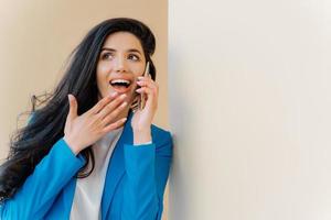 positiva donna d'affari apre la bocca per l'interesse, tocca il mento, guarda da parte, affascinata dalle voci, ha una conversazione telefonica con un caro amico, spettegola sulle ultime notizie, indossa un costume blu formale foto