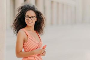 contenta ragazza millenaria con i capelli ricci, indossa un abito rosa, occhiali da vista, concentrata in disparte con un sorriso, sta all'aperto, aspetta una chiamata, gode di una connessione internet ad alta velocità, aggiorna la nuova applicazione foto