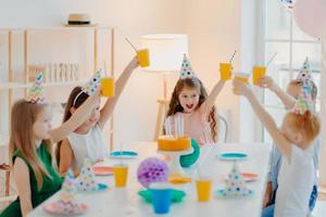 un gruppo di allegri bambini in età prescolare festeggiano il compleanno insieme, si divertono, applaudono con tazze di bevande, indossa cappelli festivi, mangia una deliziosa torta, si siede a tavola in una stanza spaziosa. bambini e concetto di festa foto