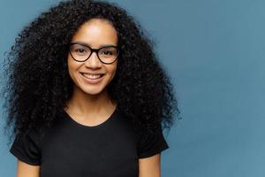 colpo alla testa di una donna dalla pelle scura dall'aspetto piacevole con un taglio di capelli afro, indossa occhiali trasparenti e una maglietta nera casual, esprime buone emozioni, si trova su sfondo blu, spazio libero per lo slogan foto