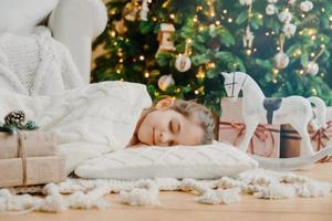 la ragazza affascinante dorme su un morbido cuscino bianco sul pavimento contro l'albero di capodanno decorato, fa sogni piacevoli, circondata da un cavallo giocattolo e scatole regalo. concetto di bambini, riposo e vacanze invernali. foto