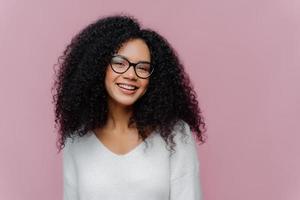 colpo alla testa di una donna adorabile e compiaciuta con l'acconciatura riccia, sorride dolcemente alla telecamera, indossa occhiali ottici e un maglione bianco casual, posa su sfondo viola. concetto di emozioni e sentimenti positivi foto