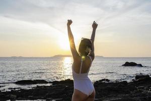 turista donna alzando le mani con il tramonto in spiaggia in vacanza. costume da bagno femminile che indossa braccia rilassanti e sollevate con granelli di sabbia sulla pelle. signora asiatica che si gode la natura in vacanza. concetto di libertà foto