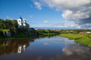 una vecchia chiesa ortodossa e un villaggio sulla riva del fiume. sole, cielo blu e nuvole nere. foto