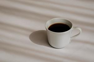 una tazza di caffè nero mette su un lenzuolo bianco con l'ombra della tenda al mattino. concetto di relax a casa. foto