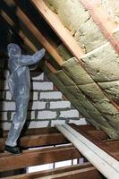 un uomo in tuta protettiva mette lana minerale tra le travi del tetto della casa per riscaldarsi dal freddo foto