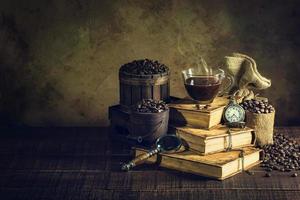 caffè in tazza di vetro su libri antichi e orologio vintage su pavimento in legno invecchiato foto