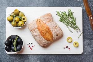 ciabatta italiana con olive e rosmarino su un tagliere. sfondo di cemento scuro. foto