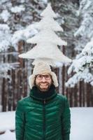 ritratto verticale di allegro uomo barbuto si diverte da solo nella foresta invernale, mantiene un abete artificiale, posa all'aperto, ammira il gelido clima nevoso, esprime positività ed emozioni piacevoli foto