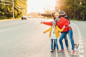 il ritratto della madre di famiglia, del padre e della figlia piccola camminano con l'animale domestico, stanno sulla strada, alzano i pollici come segno di ok, esprimono approvazione o accordo, sono di buon umore dopo aver trascorso del tempo all'aria aperta foto