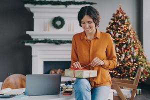sorridente felice femmina italiana che lega un nastro rosso sul regalo di Natale mentre è seduto a casa in ufficio foto