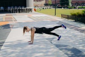 la giovane donna sportiva snella fa esercizio di fasciame alza la gamba dimostra la sua determinazione indossa abiti sportivi e scarpe da ginnastica si allena all'aperto ascolta musica negli auricolari. concetto di stile di vita sano foto