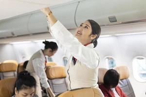 assistente di volo femminile asiatica che chiude il coperchio del bagagliaio per il bagaglio a mano dopo che i passeggeri si sono seduti e si preparano a decollare foto