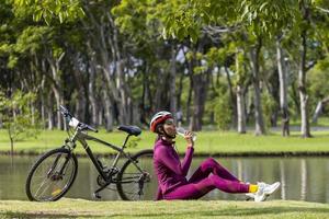 donna asiatica transgender in abbigliamento sportivo che si riposa e beve acqua per la reidratazione dopo aver pedalato con la bicicletta sportiva intorno al parco pubblico in riva al lago foto