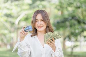 sorridente giovane libero professionista che mostra la carta di credito e tiene banconote di denaro sfocato sfondo verde, donna felice che tiene e mostra banconote di denaro foto