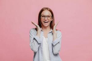 studio di una donna gioiosa eccitata vestita con una camicia a righe, allarga le mani vicino al viso, indossa occhiali ottici, modella su sfondo rosa, non si aspetta di notare qualcosa di piacevole e imprevedibile foto