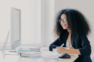 il manager professionista di una donna dai capelli ricci fa un rapporto, si concentra sullo schermo, annota le informazioni, indossa occhiali e abito formale, prende appunti sull'idea per la pianificazione della partenza, posa all'interno dell'ufficio. foto