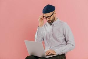 copywriter maschio caucasico vestito con abiti eleganti, lavora come freelance, digita informazioni sul computer portatile, connesso a Internet wireless, ha un aspetto attento, isolato su sfondo rosa foto