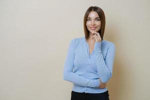 mezza lunghezza di una donna piuttosto snella vestita con un maglione blu casual, tiene la mano sotto il mento, gode di una pelle liscia e sana, pronta per il lavoro, isolata su sfondo beige, spazio vuoto a parte foto