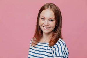 inquadratura laterale di una donna dai capelli castani dall'aspetto piacevole e felice con capelli lisci, pelle sana, sorriso piacevole, vestita con un maglione a righe casual, isolata su sfondo rosa. concetto di emozioni foto