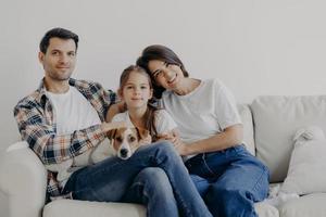 ritratto di affettuosa famiglia coccola e si siedono insieme sul divano in soggiorno, cambiano casa, hanno espressioni felici. padre, madre, figlia e cane posano per fare ritratti, trascorrono del tempo foto