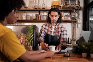 di fronte al primo piano, un giovane barista maschio tailandese dai capelli lunghi lavora servendo una tazza di caffè a una cliente afroamericana al bancone della caffetteria, un buon lavoro di servizio e una piccola imprenditrice. foto