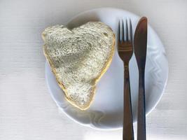 vista dall'alto da vicino, pagnotta fatta in casa a forma di cuore, fetta di pane, piatto bianco con forchetta e coltello sulla tovaglia bianca foto