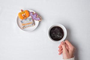 mano della donna che tiene la tazza di caffè durante il consumo di biscotti divertenti di halloween. buon giorno di halloween, trucco o minaccia, ciao ottobre, autunno autunno, tradizionale, festa e concetto di vacanza foto