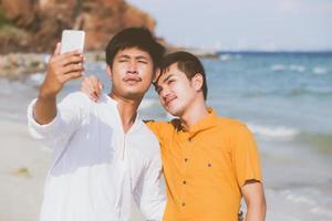 ritratto gay giovane coppia sorridente che scatta una foto selfie insieme al telefono cellulare intelligente in spiaggia, amante omosessuale lgbt in vacanza al mare, due uomini che vanno in viaggio, concetto di vacanza.