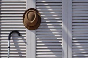 luce solare e ombra sulla superficie del cappello fedora con bastone da passeggio in metallo su sfondo bianco muro pieghevole in legno in stile vintage tono foto