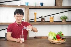 il giovane asiatico con una maglietta rossa casual si diverte a fare colazione, bevendo latte. giovane che cucina cibo nella stanza della cucina in stile loft foto