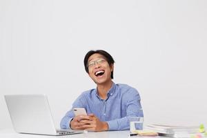 allegro attraente asiatico giovane uomo d'affari in occhiali e camicia blu utilizzando il telefono cellulare lavorando con il computer portatile e ridendo su sfondo bianco foto