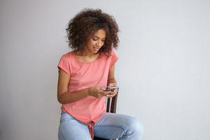 attraente giovane donna dalla pelle scura con capelli ricci castani seduta su una sedia e con in mano uno smartphone, chiacchierando con gli amici, posando su un muro bianco con una t-shirt rosa foto