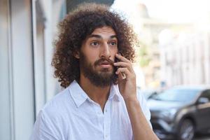 sorpreso bel ragazzo barbuto con capelli ricci castani che cammina per strada con il cellulare in mano, corrugando la fronte e alzando le sopracciglia foto