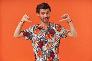 giovane attraente fiducioso con stoppia in camicia hawaiana in piedi e puntato su se stesso con i pollici su entrambe le mani su sfondo arancione foto