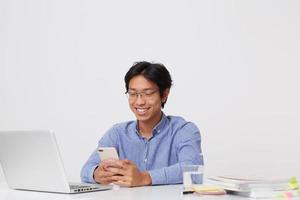 sorridente bel giovane uomo d'affari asiatico in occhiali e camicia blu utilizzando il telefono cellulare e laptop seduto al tavolo su sfondo bianco foto