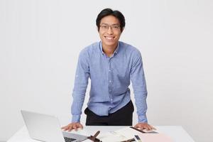 felice fiducioso asiatico giovane uomo d'affari in bicchieri con auricolari guardando la fotocamera in piedi vicino al tavolo con laptop isolato su sfondo bianco foto