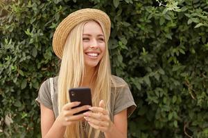 sorridente giovane bella donna che guarda da parte felicemente, indossa abiti casual e cappello di paglia, posa su un giardino verde, tenendo in mano lo smartphone foto