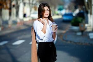 giovane donna alla moda che beve il tè in una strada della città. ragazza europea hipster con bicchiere di carta latte. splendida giovane donna con una tazza di caffè in una strada di città. pausa caffè. caffè da portar via. foto