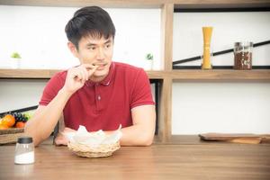 il giovane asiatico con una maglietta rossa casual si diverte a fare colazione, mangiando patatine fritte. giovane che cucina cibo nella stanza della cucina in stile loft foto