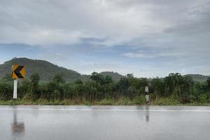 vista orizzontale della strada asfaltata bagnata in Thailandia. ambiente di tempo piovoso. e lo sfondo della piantagione di manioca e delle montagne verdi. sotto il cielo azzurro e nuvole scure. con segnale stradale curvo. foto