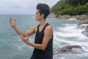 giovane in abbigliamento sportivo che fa arte marziale, qigong, tai chi sulla roccia al mare, concetto di salute e meditazione foto