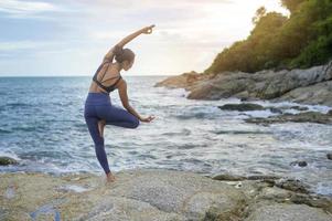 giovane donna asiatica in abbigliamento sportivo che fa yoga sulla roccia al mare, concetto di salute e meditazione foto